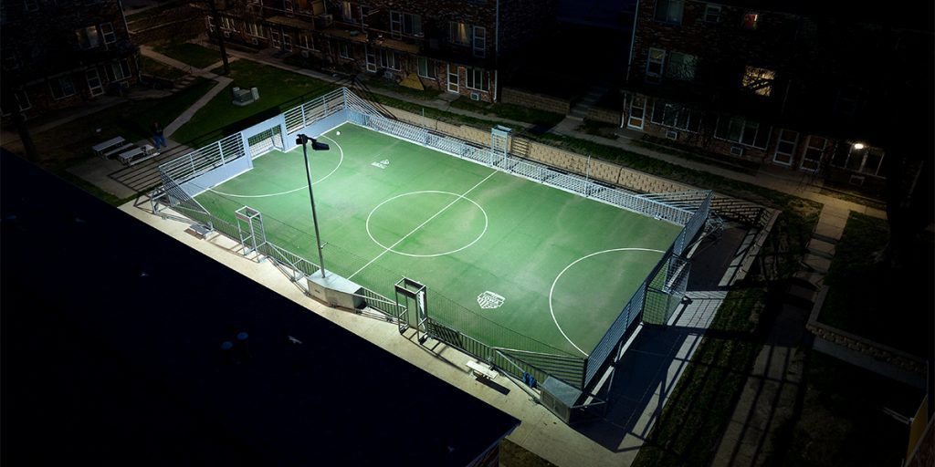 Musco Mini-Pitch System™ Modular Sports Solution situated between four of the brick apartment buildings that make up the Oakridge Neighborhood. Mini-pitch is lit at night with two factory-aimed TLC for LED fixtures, limiting spill and glared