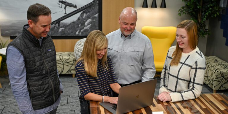 Two male and two female Musco employees look at a laptop on a wood table.