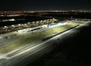 Sideview of Trinity Rail Equipment Maintenance Facility in Dallas-Ft. Worth lit with new LED rail yard lighting with commuter train passing under light poles in the foreground.