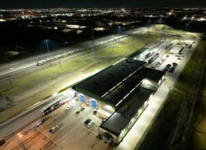 Entrance of Trinity Rail Equipment Maintenance Facility in Dallas-Ft. Worth lit with new LED rail yard lighting with commuter train passing under light poles in the background.