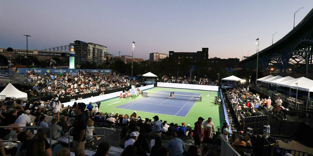 Blue and green tennis court at Tennis in the Land surrounded by people in the stands and lit at sunset with Musco temporary lighting system with LED fixtures.