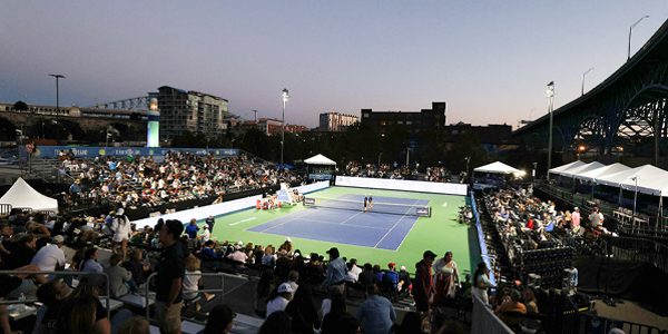 Blue and green tennis court at Tennis in the Land surrounded by people in the stands and lit at sunset with Musco temporary lighting system with LED fixtures.