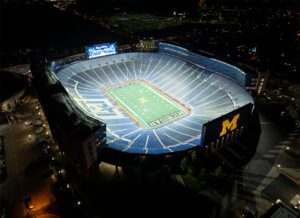 Overhead view of University of Michigan's Michigan Stadium stands and football field lit at night.