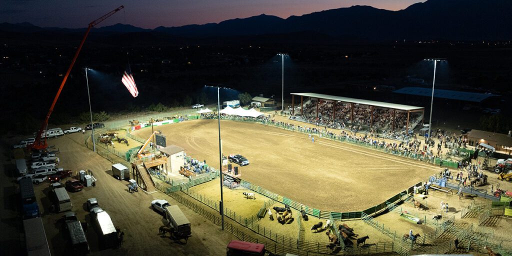 Rodeo arena lights turn on at Douglas County Fairgrounds as the sunsets. An American flag is flying from a crane on the left while spectators gather in the stands.